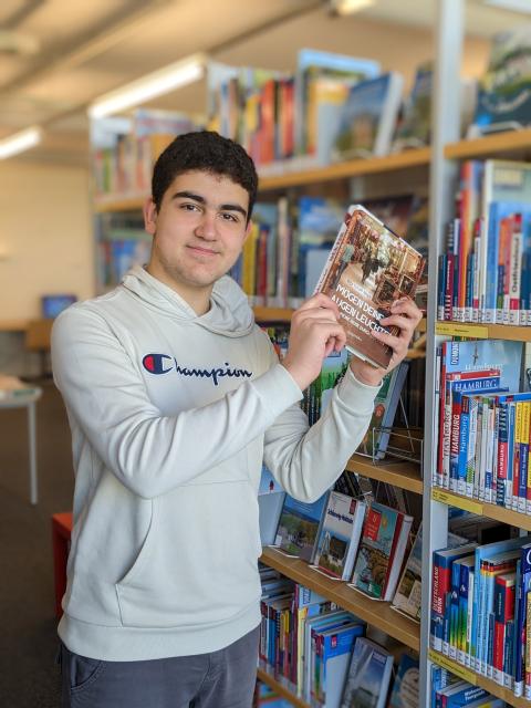 Ein Junger Mann steht vor einem Bücherregal mit einem Buch in der Hand